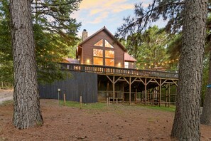 Surrounded by mature trees, this cabin features a gabled roof and a large window, with a wooden deck extending over a dirt area.