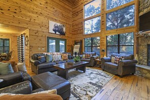 Relax in this spacious living room with a central coffee table, leather sofas, and armchairs, surrounded by large windows offering a view of the surrounding trees and a stone fireplace.