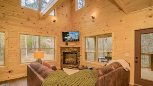 A cozy cabin interior with a stone fireplace, large windows, and a comfortable seating area. - 🌅 Bear Valley Splash 🍃 Indoor Pool, Creekside Yard, Hot Tub & Games (Sevierville)