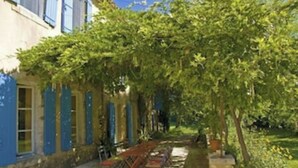 Une cour avec une pergola, des sièges d’extérieur et un bâtiment aux volets bleus.