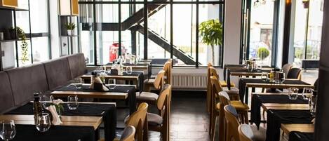 A modern restaurant with high ceilings, large windows, and a staircase leading to an upper level. Tables are arranged for dining, and there are plants and hanging lights.