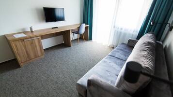 Una habitación de hotel con un escritorio de madera, un televisor, un sofá gris y una ventana con cortinas.