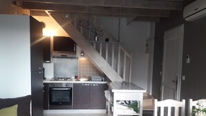 Ett kompakt kök med en gasspis, ugn och ett litet matbord med stolar. En trappa leder upp till en övre våning, och en soffa syns i förgrunden.