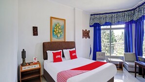 A hotel room with a large bed, a nightstand with a lamp, a painting on the wall, and a view of the outdoors through the window.