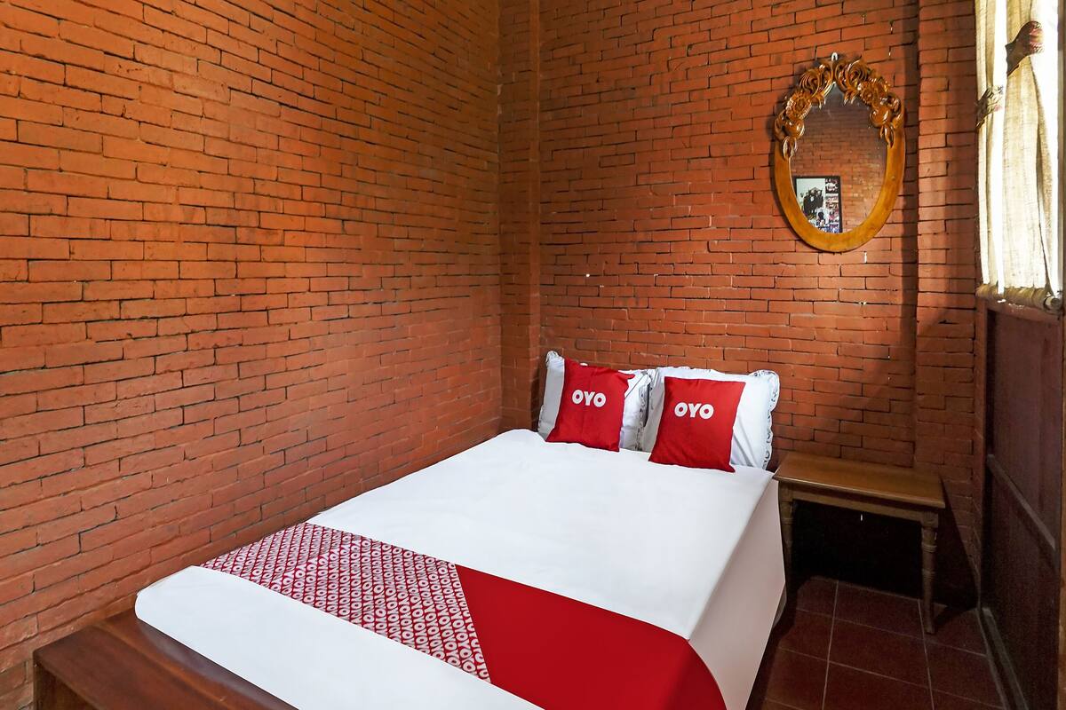 A single bed with a white and red bedspread, two pillows with the word "OYO" on them, a wooden bench, a mirror on the wall, and a brick wall.