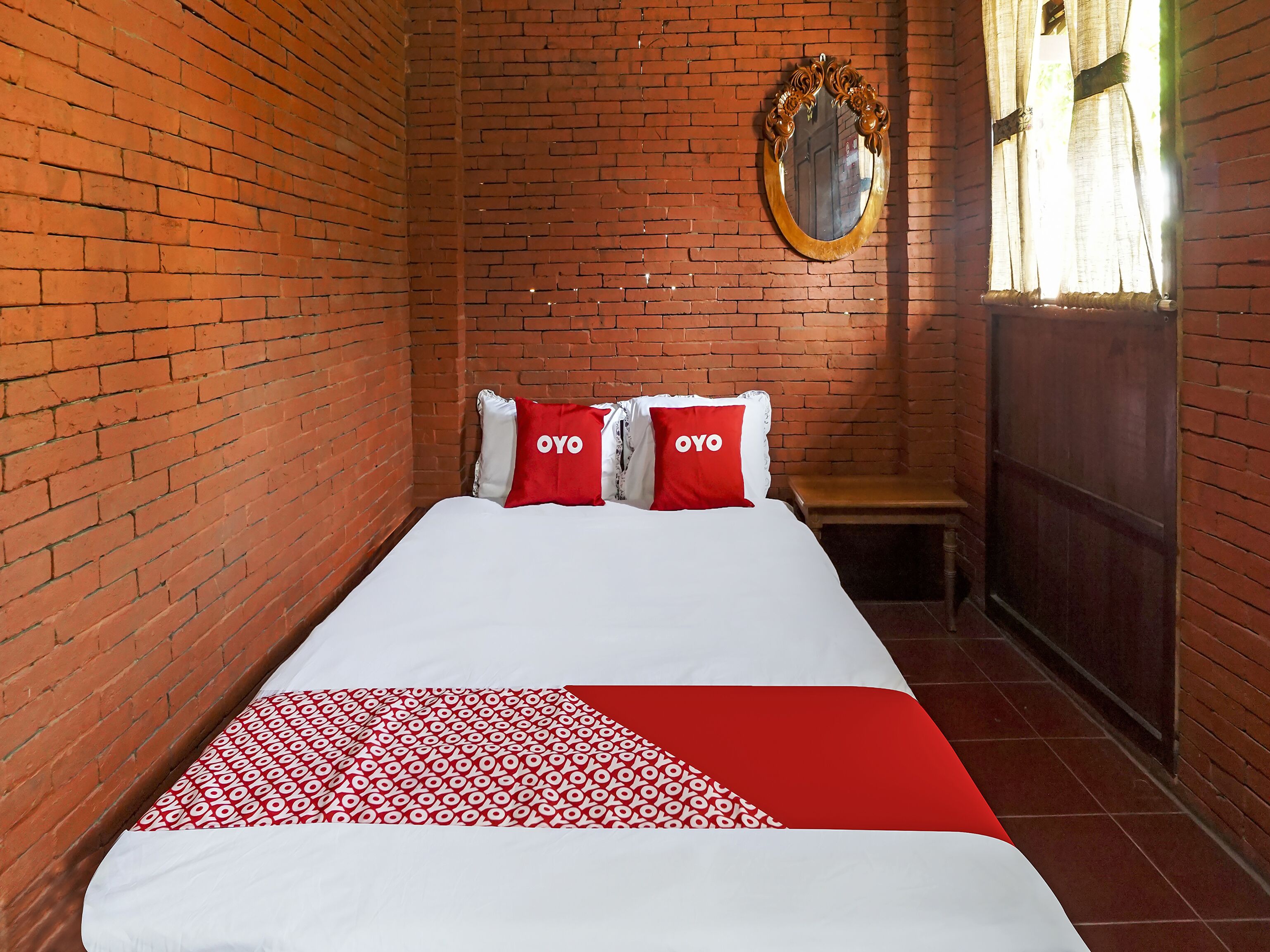 A narrow room with a brick wall, a bed with white bedding and red pillows, a wooden bench, and a mirror on the wall.