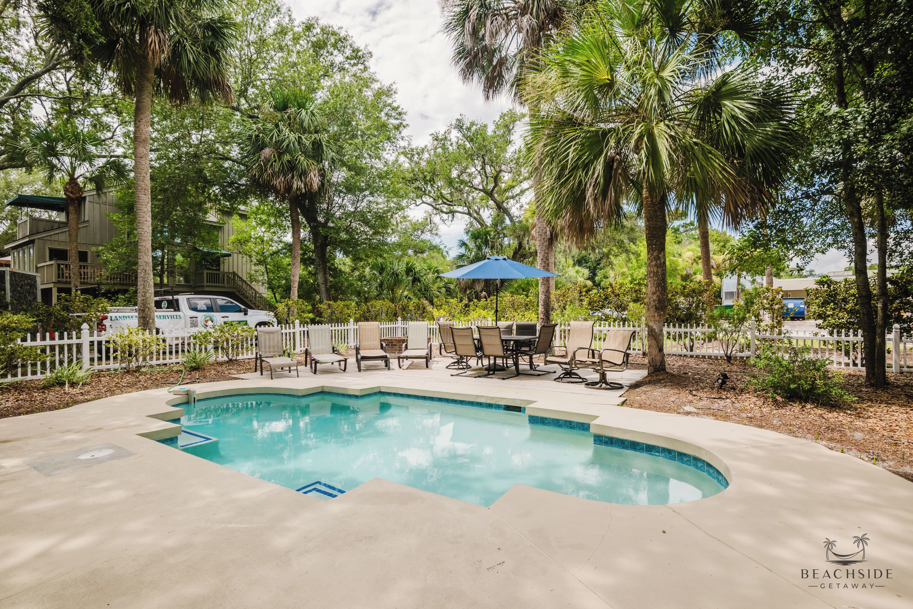 A swimming pool with a white fence, lounge chairs, and a palm tree.