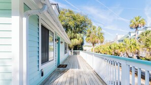 A wooden deck with a white railing, a covered porch, and a view of palm trees and other greenery.