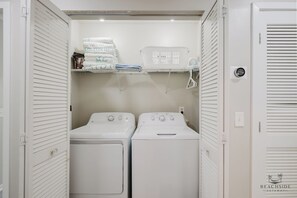 Une buanderie équipée de deux machines à laver à chargement frontal, d’une étagère où sont pliées les serviettes et d’un thermostat fixé au mur.