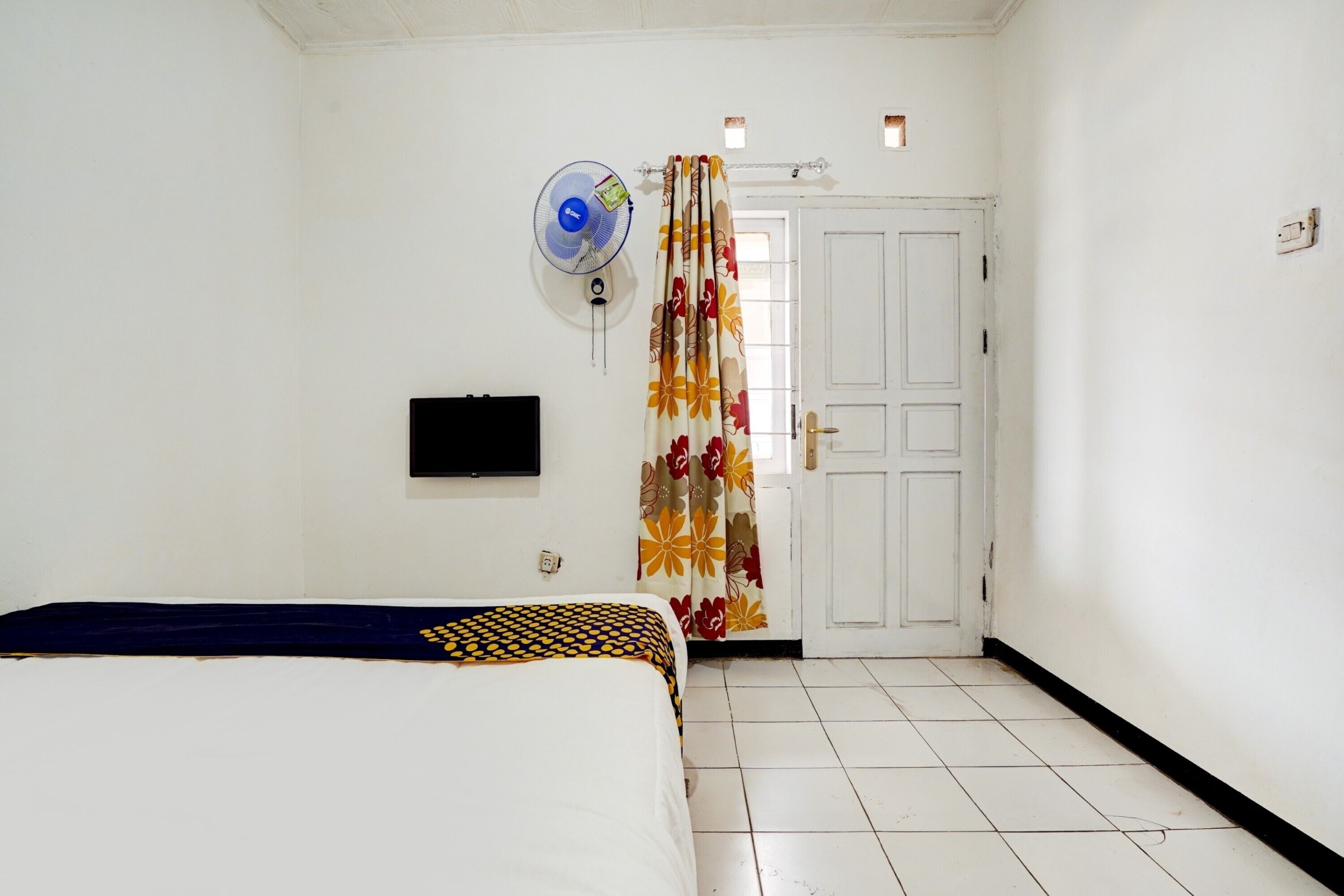 Una stanza con un letto, una TV a muro, una ventola a soffitto e una porta con tenda a fiori.