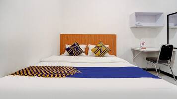 Una cama bien hecha con una colcha azul y amarilla de estampado, dos almohadas con diseños geométricos, un cabecero de madera, una mesita de noche blanca con un estante pequeño y una silla negra.
