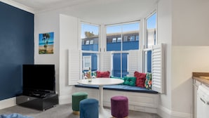 A room with a bay window, a flat-screen TV, a round table, and colorful ottomans. - Caldey Bay Apartment - Lovely Sea Views (Tenby)