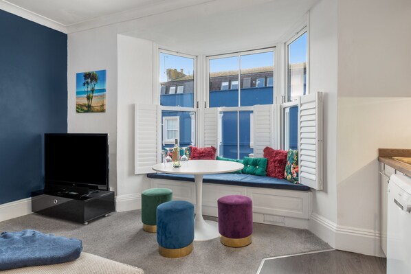 A room with a bay window, a flat-screen TV, a round table, and colorful ottomans.