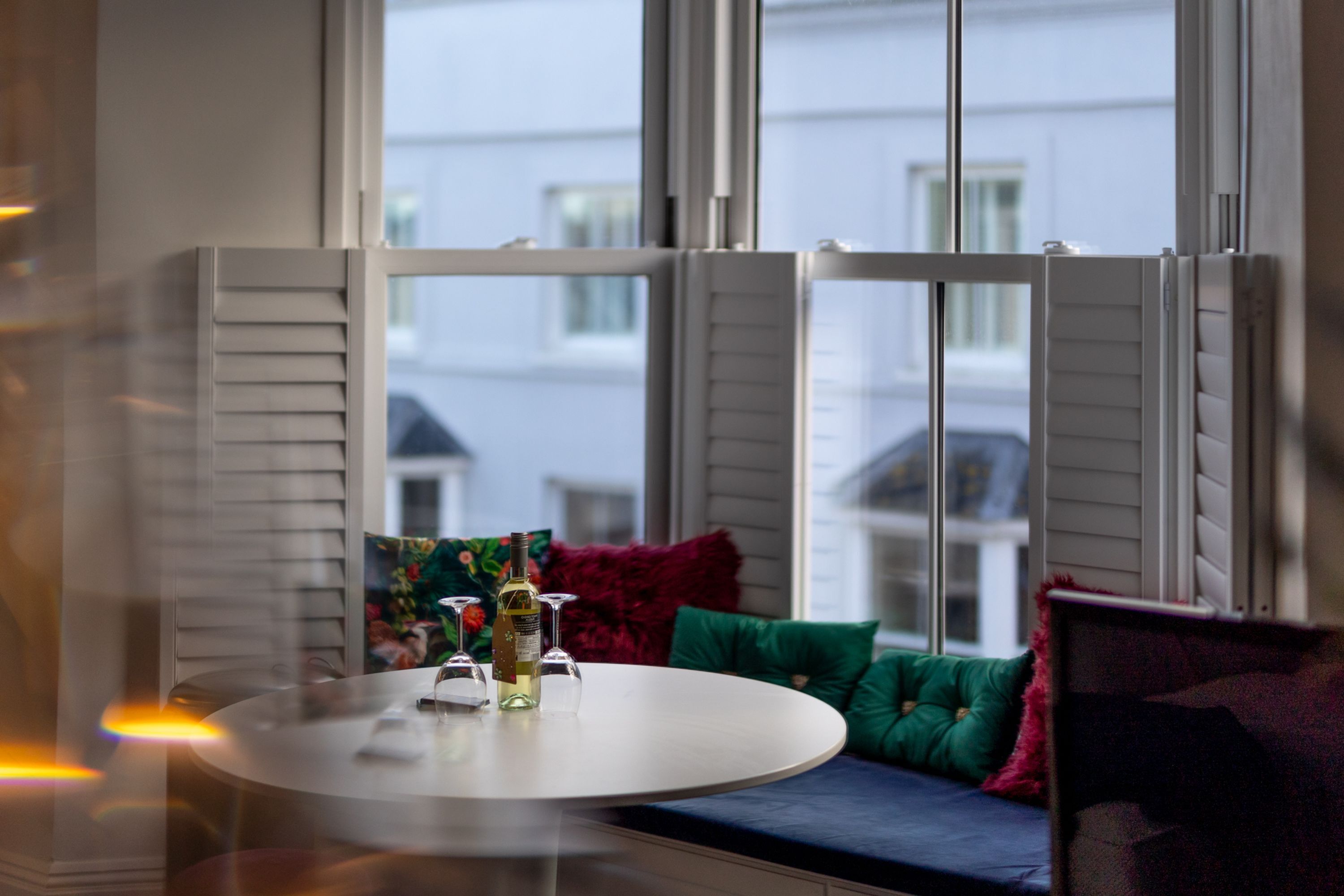 Une pièce avec une table ronde, une bouteille de vin et des verres. Il y a une fenêtre avec des volets et une vue sur les bâtiments à l’extérieur.
