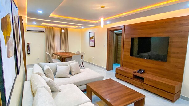 A modern living room with a large sectional sofa, a wooden coffee table, and a flat-screen TV mounted on a wooden wall. - PLUSH HOTEL (Abuja)