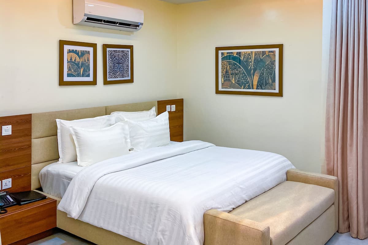 Quarto de hotel moderno com cama, mesa de cabeceira e um banco. Há unidades de ar condicionado na parede, obras de arte emolduradas e um lustre.