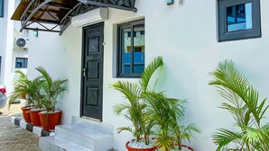 A building with a white exterior, a black door, and a small window. There are three potted plants on the ground in front of the building. - PLUSH HOTEL (Abuja)