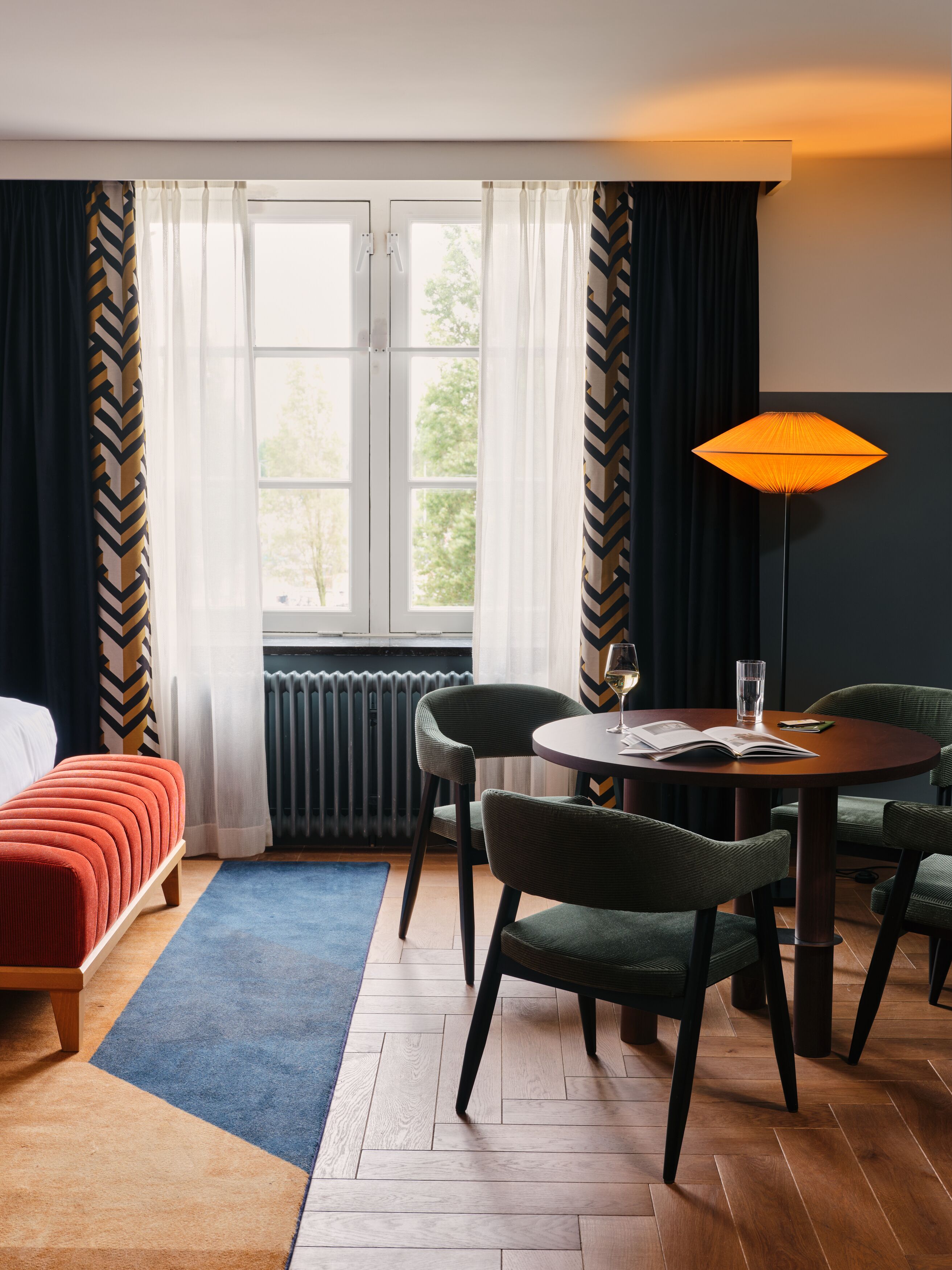 Habitación de hotel con cama, una mesa redonda con sillas, una lámpara de pie y vistas a árboles por la ventana.