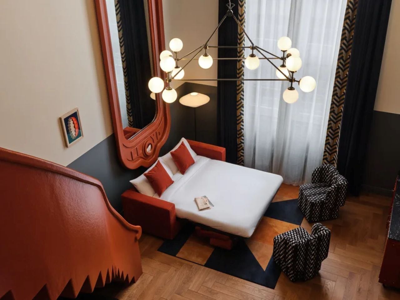 A hotel room with a bed, a mirror, a chandelier, and a wooden floor.