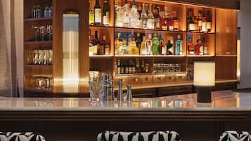 Eine Bar mit einer Holztheke, Regalen voller Spirituosenflaschen, Gläsern und einem Schiffsmodell. Drei gepolsterte Barhocker mit geometrischem Muster sind vorhanden.