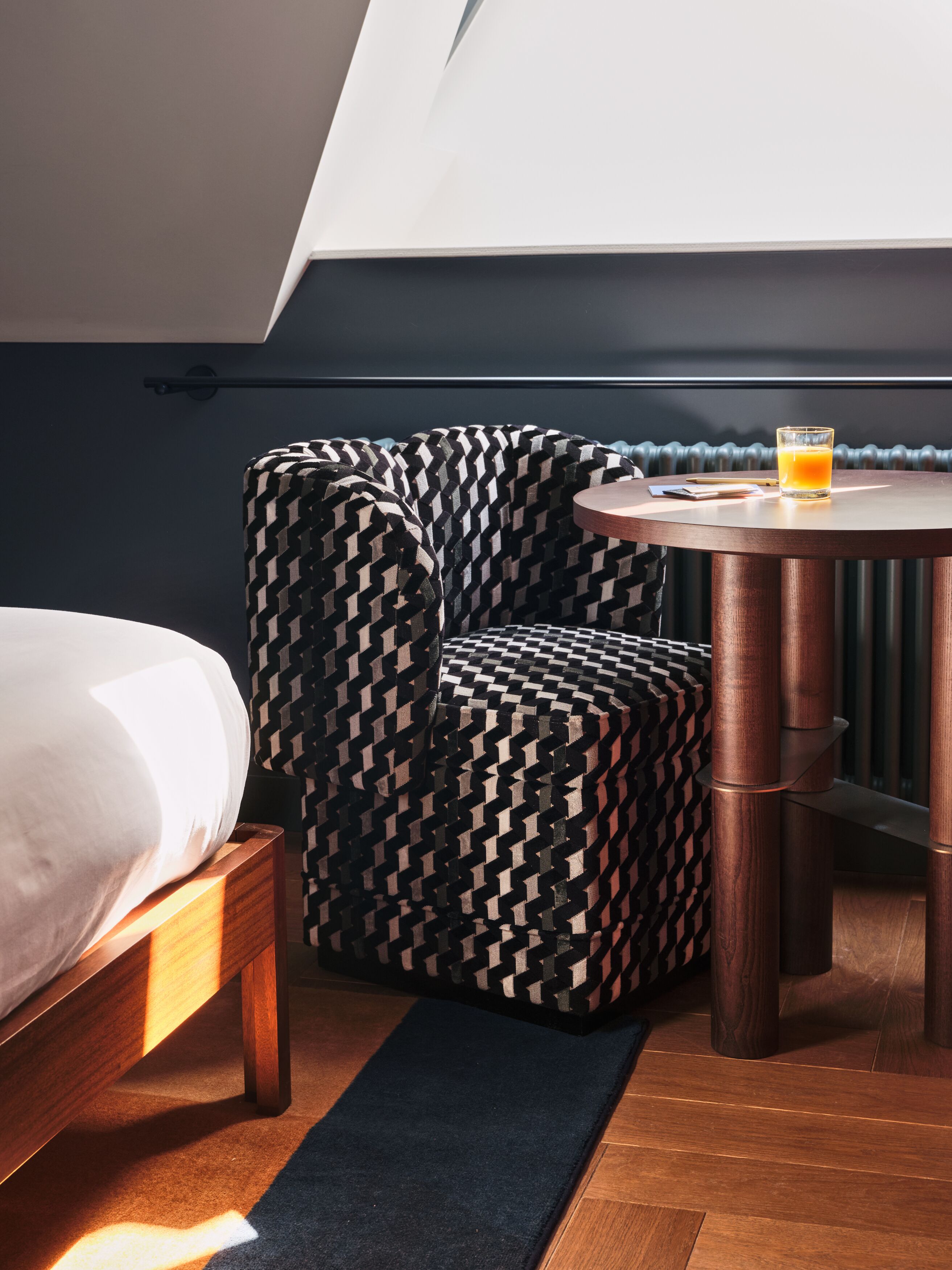 A modern hotel room with a bed, a patterned armchair, a small round table with a glass of orange juice, and a radiator.
