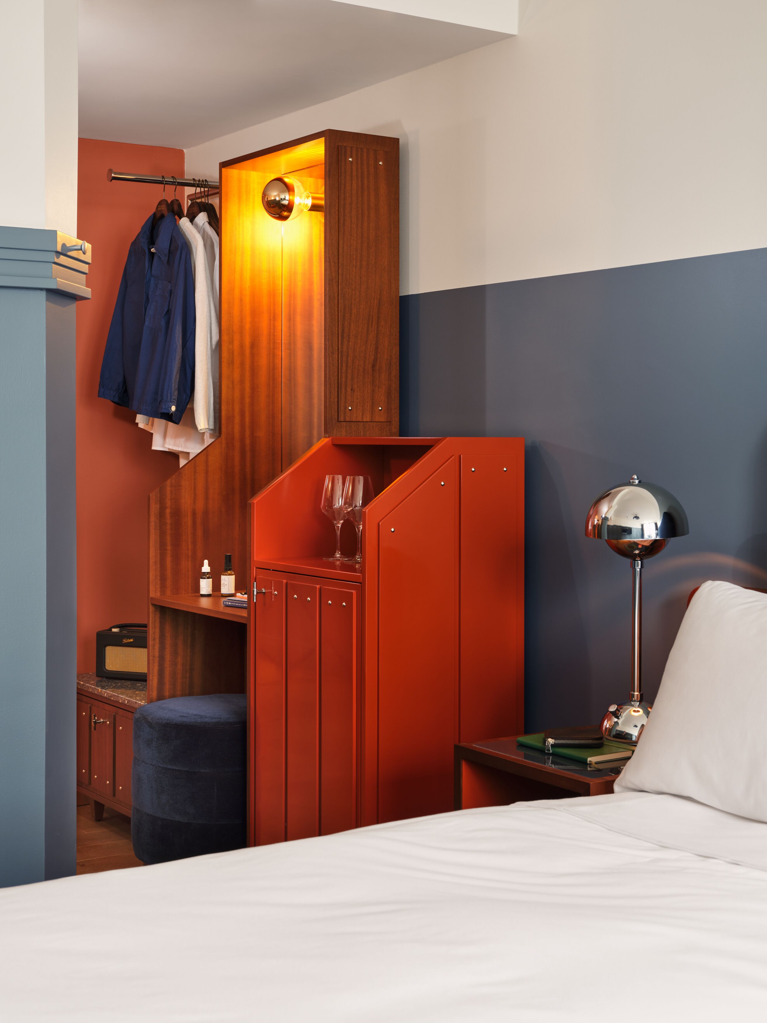 A hotel room with a bed, a nightstand, a lamp, a wardrobe, and a seating area with a chair and a small table.