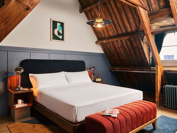 A bedroom with a wooden ceiling, a bed with white linens, a red ottoman, and a framed artwork on the wall.