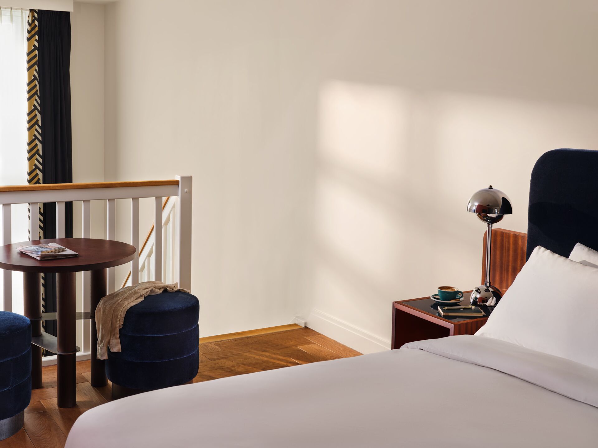 Een hotelkamer met een bed, een klein tafeltje met een tijdschrift, een stoel, een lamp en een raam met gordijnen.