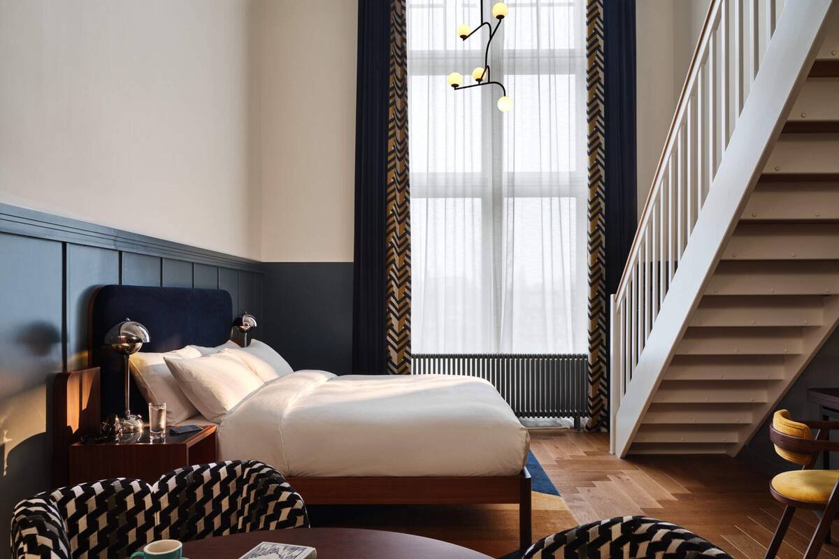 A hotel room with a bed, a bedside table with a lamp, a chair, a table with a book and a cup, and a staircase.