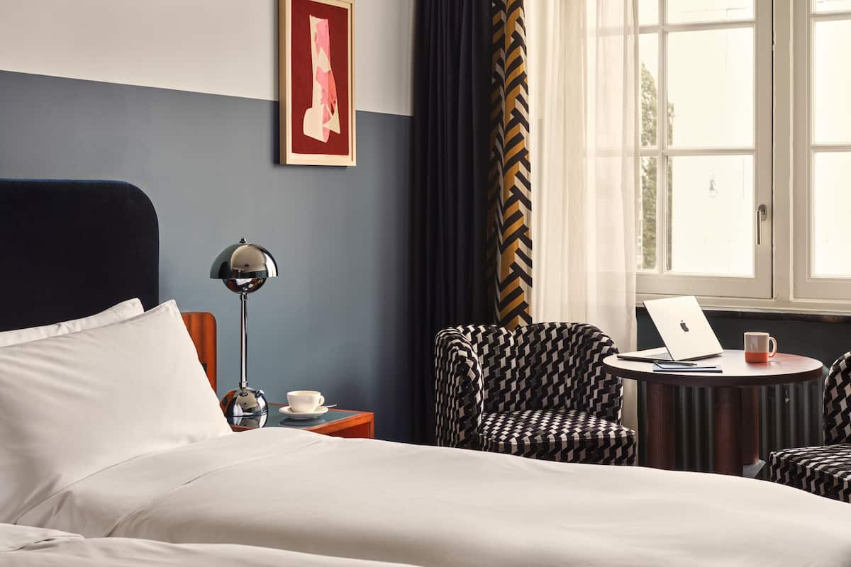 A hotel room with a bed, a desk with a laptop, a chair, a lamp, and a framed picture on the wall.