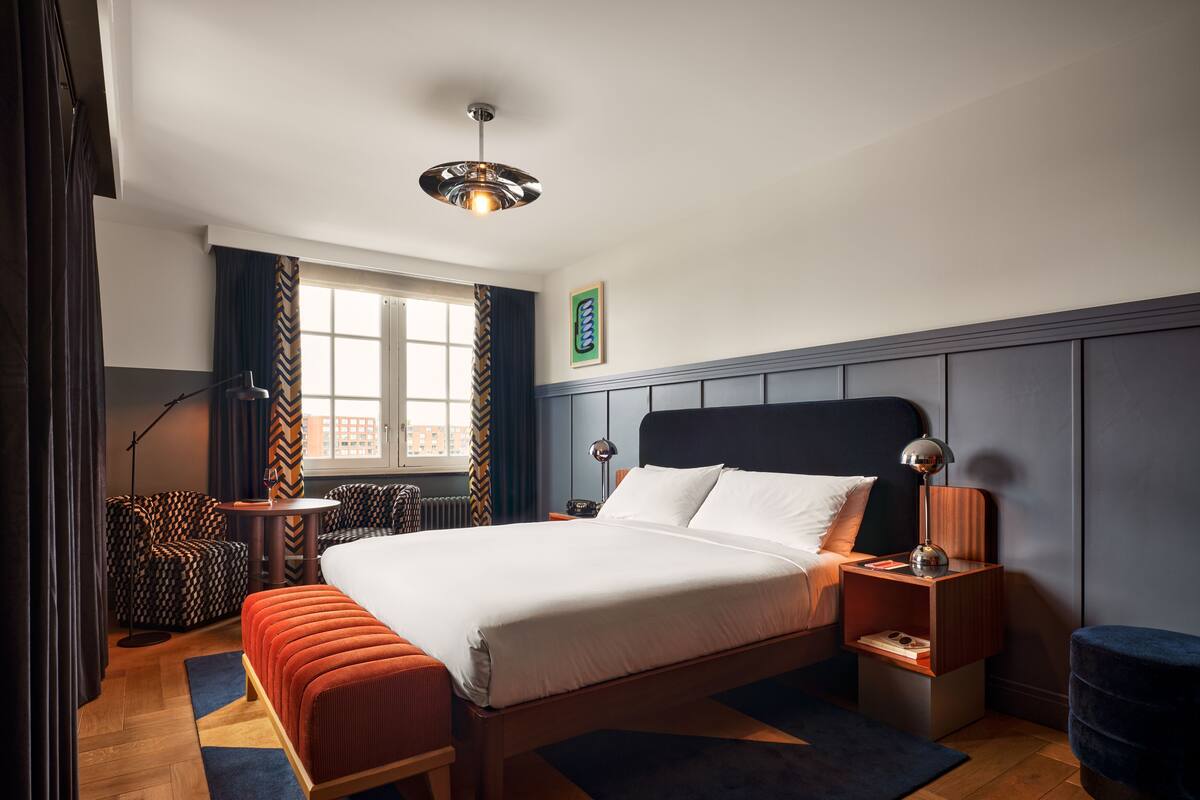 A modern hotel room with a large bed, a wooden floor, a desk, a chair, and a window with curtains.