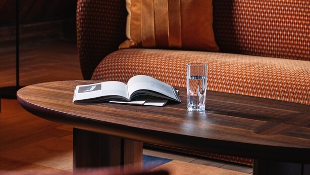 Una habitación acogedora con techo de madera, un sofá con cojines estampados, una mesa redonda de madera con un libro y un vaso de agua, y un televisor en la pared.