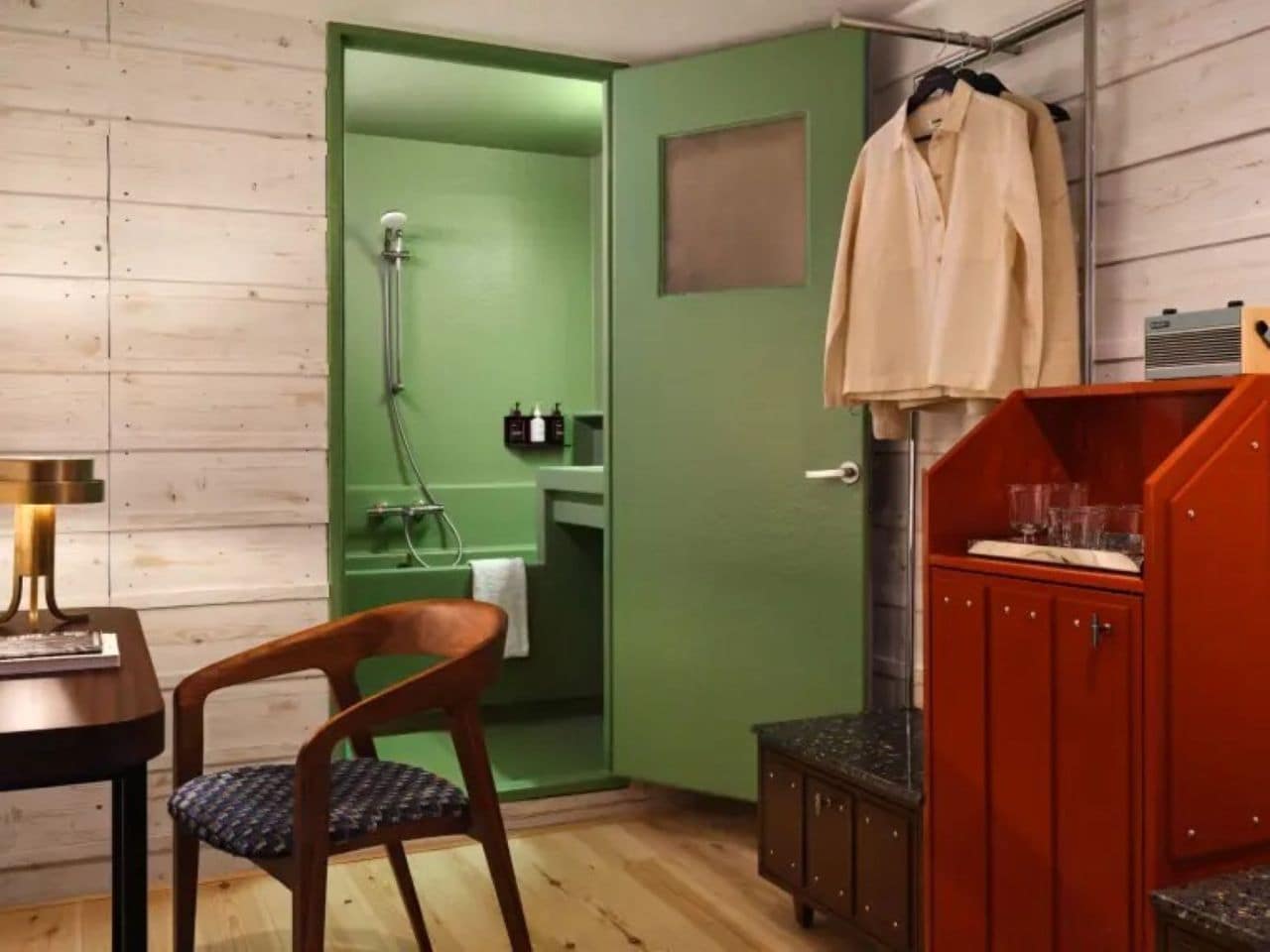 A compact room with a wooden floor, a green door leading to a bathroom, a wooden chair, a small table with a lamp, and a red cabinet.