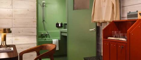 A compact room with a wooden floor, a green door leading to a bathroom, a wooden chair, a small table with a lamp, and a red cabinet.
