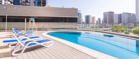 A rooftop swimming pool with blue lounge chairs and a city skyline in the background.