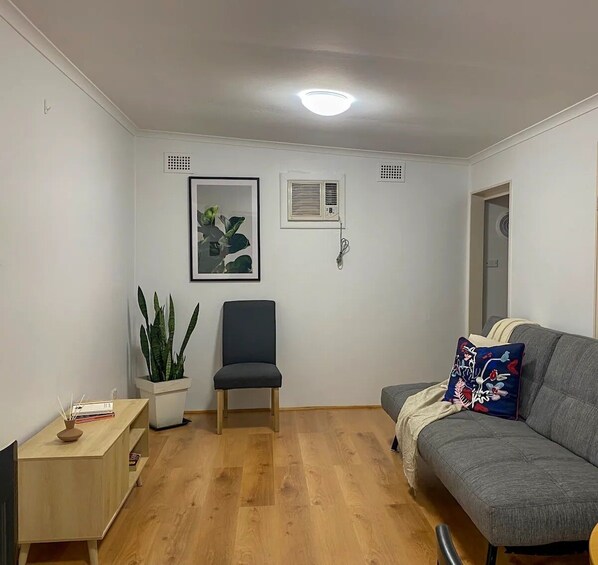 A living room with a grey sofa, a black chair, a wooden cabinet, a potted plant, and a framed picture on the wall.