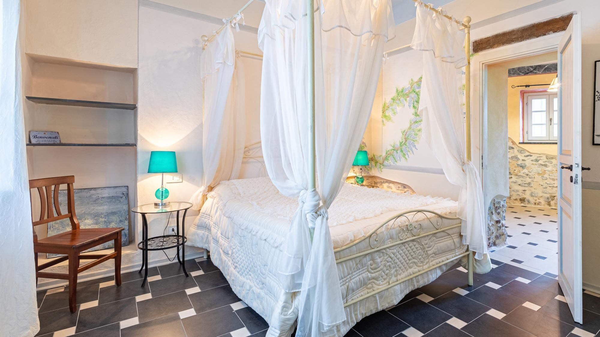 A four-poster canopy bed with a quilted duvet, a small round table with a lamp, a wooden chair, and a checkered floor.