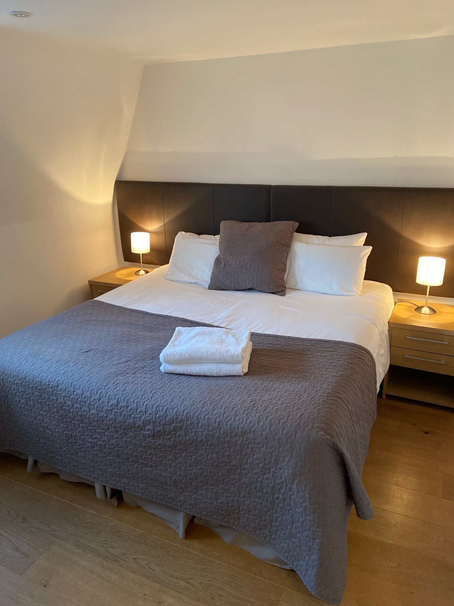 Ein ordentlich gemachtes Bett mit grauer Decke, zwei Kissen und einem gefalteten Handtuch. An beiden Seiten des Bettes befinden sich Nachttische mit Lampen.