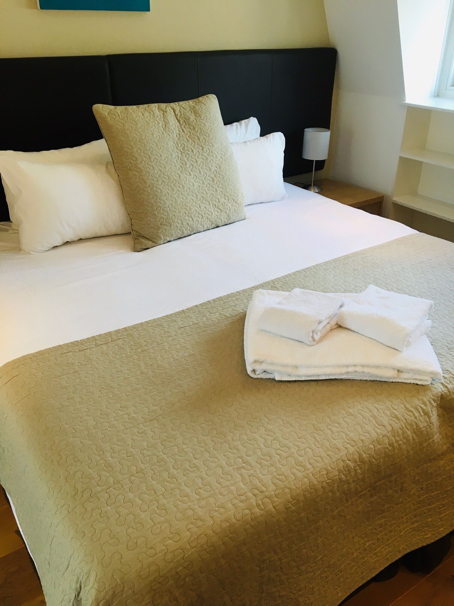 Ein ordentlich bezogenes Bett mit einer beige farbenen Steudecke, weißen Kissen und einem gefalteten weißen Handtuch.