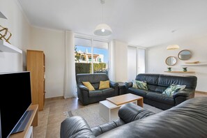 A modern living room with a flat-screen TV, leather sofas, a wooden coffee table, and a large window offering a view of buildings outside.