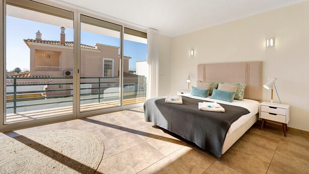Une chambre dotée d’une grande porte-fenêtre coulissante, d’un lit recouvert d’une couverture à carreaux gris et blancs, d’une table de chevet avec une lampe, et offrant une vue sur un bâtiment dont le toit est recouvert de tuiles.