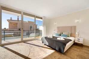 A bedroom with a large sliding glass door, a bed with a grey and white checkered bedspread, a nightstand with a lamp, and a view of a building with a tiled roof.