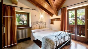 Une chambre avec un lit, une fenêtre, un radiateur, des poutres en bois au plafond et une vue sur la verdure à l’extérieur.