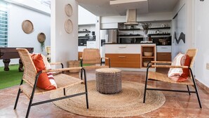 A modern living room with wicker furniture, a round ottoman, and a kitchen in the background.