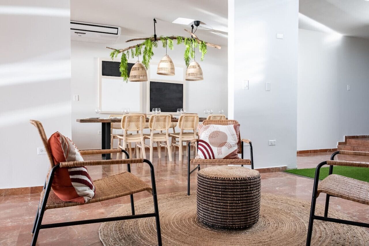 Un salon moderne avec un coin repas, comprenant des meubles en osier, un pouf rond en fibres tressées et un luminaire suspendu au plafond agrémenté de plantes retombantes.