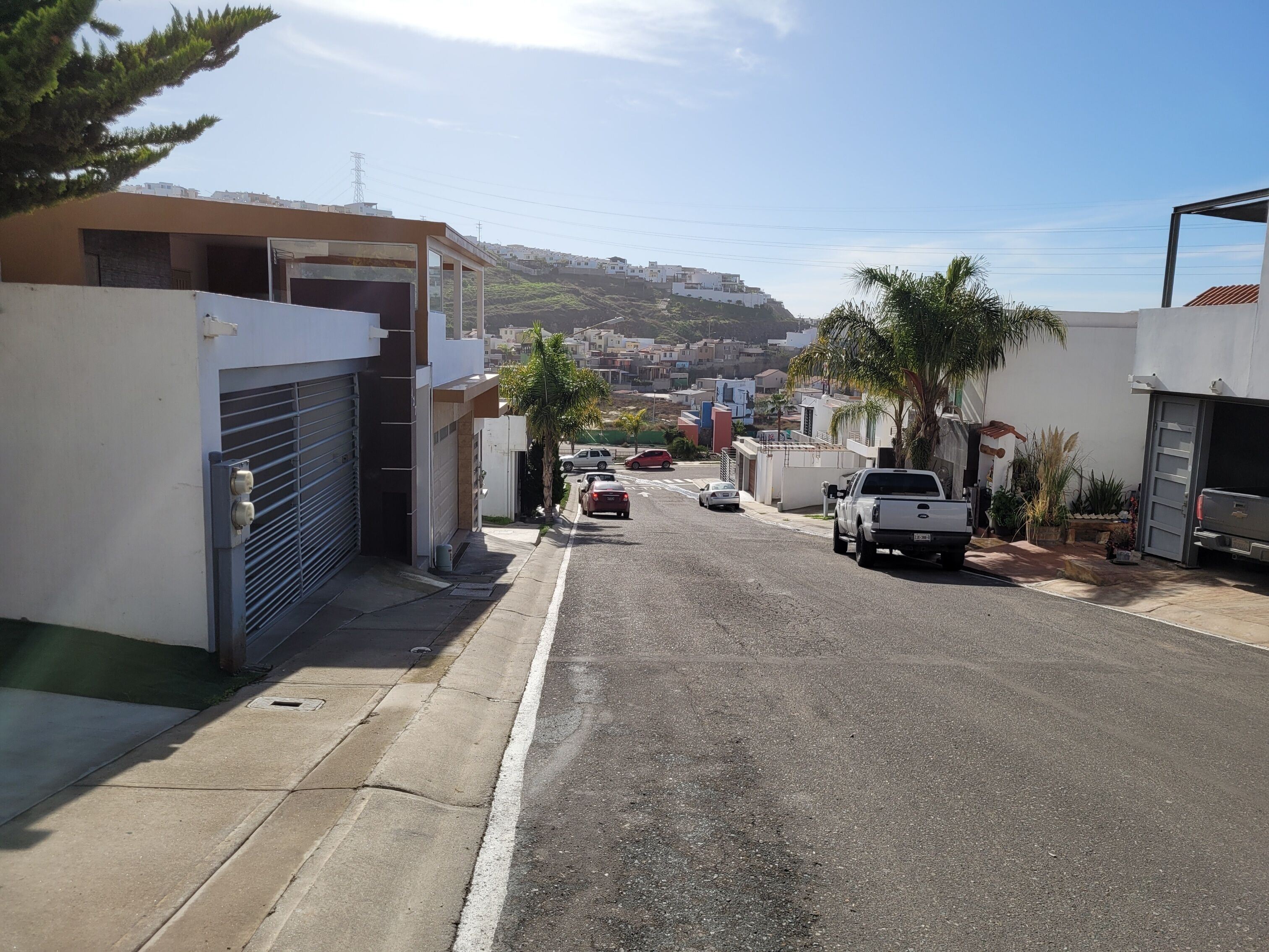 A residential street with houses, parked cars, and a clear view of a hilly town in the distance.