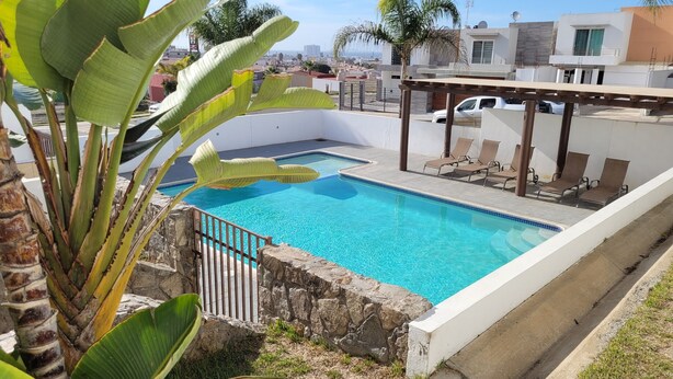 Une piscine bordée d’un mur en pierre et entourée d’une clôture en métal, avec des chaises longues et une pergola à proximité.