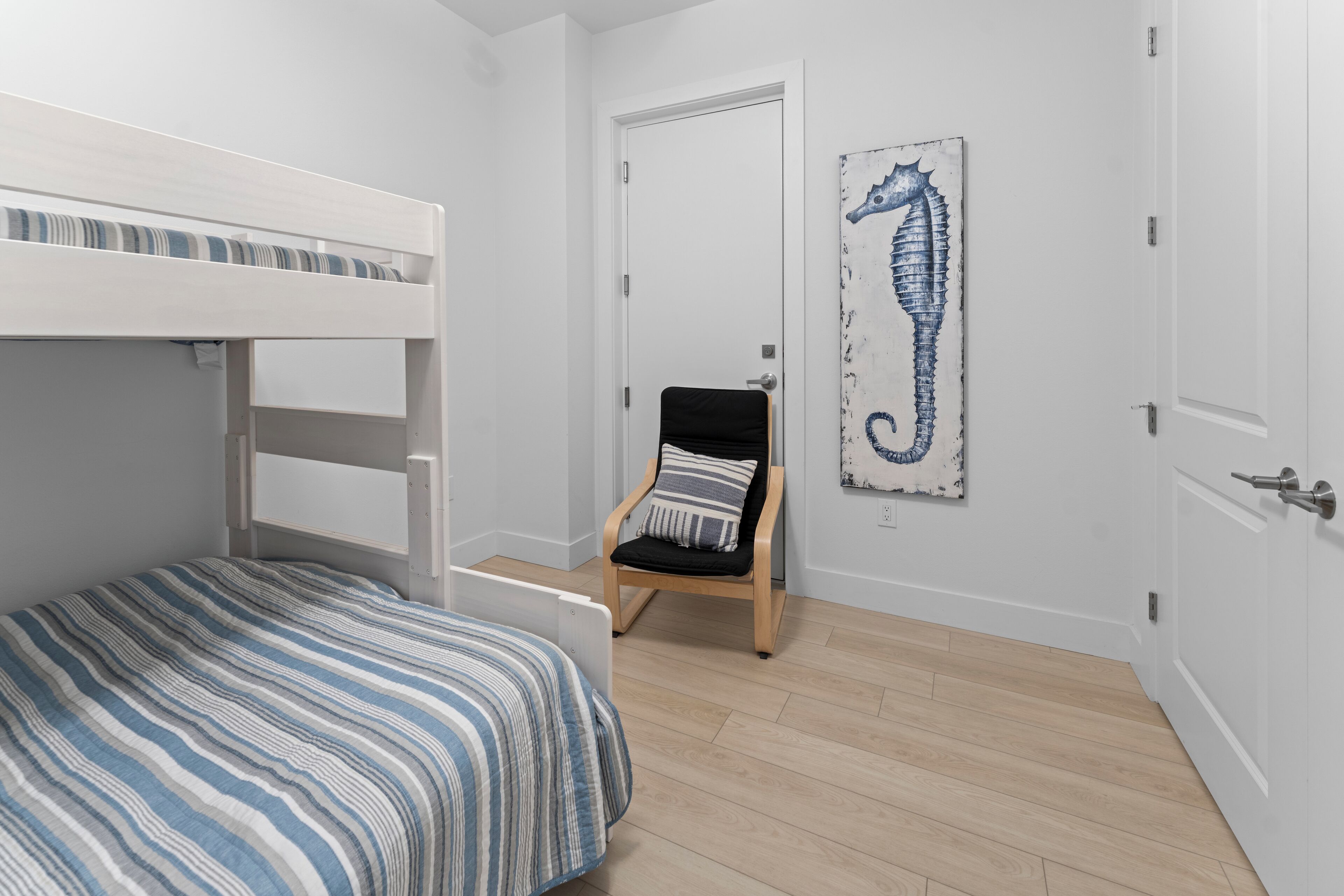 Un lit superposé avec une couvre-lit à rayures, une chaise en bois avec un coussin et une œuvre d’art représentant un hippocampe accrochée au mur.