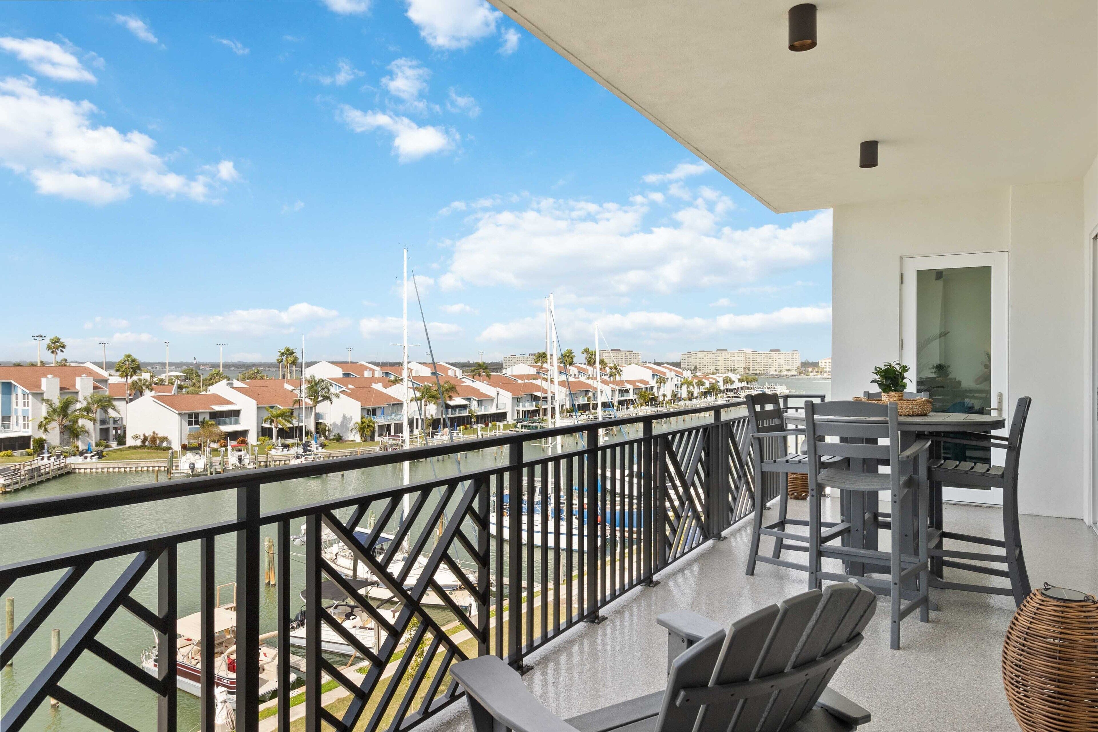 A balcony with a view of a marina, a dining area with chairs and a table, and a cityscape in the background.