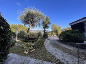 Un chemin de pierre mène à un espace aménagé avec des meubles de jardin, le tout entouré d’une végétation luxuriante et d’un grand arbre.